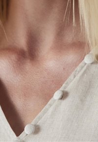 Vêtement en lin avec un décolleté en V, présentant de petits boutons en tissu le long du bord. Couleur gris clair avec une surface texturée.