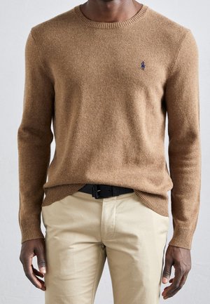 Homme portant un pull à manches longues couleur chameau avec un petit logo sombre sur la poitrine et un pantalon beige avec une ceinture noire.