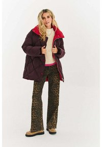 Fioletowa pikowana kurtka puchowa z czerwonym kołnierzem, noszona na kremowym swetrze i rozszerzanych spodniach w panterkę. Beżowe buty dopełniają stylizację.