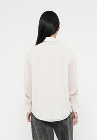 Light beige long-sleeve shirt with a soft, smooth texture, featuring a classic collar and rounded hem, worn with gray, loose-fitting pants.