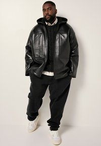 Uomo che indossa una giacca di pelle nera, un maglione di lana nero a trecce, una camicia bianca, pantaloni neri larghi e sneaker bianche, in piedi contro un muro uniforme.