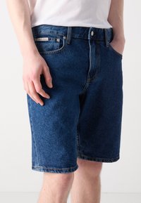 Denimshorts i mørkeblå med rett passform. Har fem lommer, belteholdere og synlig søm langs kanten og lommene.