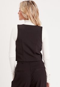 Top corto nero con design strutturato, caratterizzato da una texture liscia e cuciture posteriori. Indossato sopra una maglietta a maniche lunghe bianca a coste.