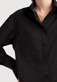 Čierna košeľa na gombíky s bielymi vertikálnymi pruhmi, vybavená náprsné vreckom a textúrovaným materiálom. Dizajn s golierom a detailmi z gombíkov.