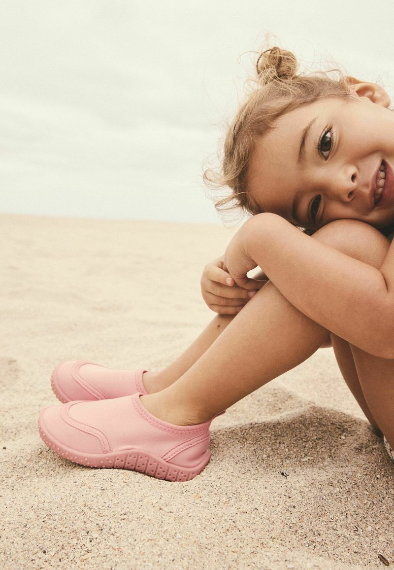 Scarpe slip-on rosa con suole imbottite, texture liscia e punta arrotondata, posizionate su una superficie di sabbia. Design semplice senza adornamenti.