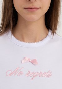 Hvid T-shirt med bølget krave, der har lyserødt broderet tekst "No regrets" og en lille satin sløjfe. Glat tekstur.