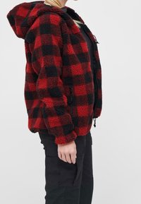 Czerwona i czarna polarowa kurtka w kratę z kapturem, zapinana na zamek błyskawiczny, z regulowanymi sznurkami i puszystą teksturą. Noszona na czarnych spodniach.