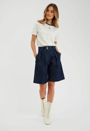T-shirt blanc avec bordures bleues et logo, associé à des culottes en denim foncé avec poches et double boutons à la taille haute. Bottines blanches à lacets.