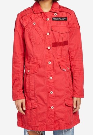 Rød langærmet jakke med knapper, flere klaplommer og syningsdetaljer, båret over revnede blå jeans.