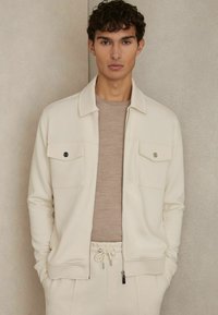 Lys cremefarvet jakke med lynlås og to brystlommer, parret med en beige skjorte. Blødt stof, afslappet pasform og minimalisk design.