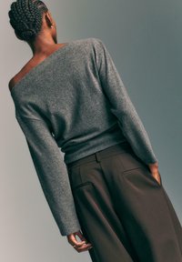 Szary, sweter odsłaniający ramiona, wykonany z miękkiego dzianiny, w połączeniu z brązowymi spodniami z wysokim stanem, które mają tylne kieszenie i luźny krój.