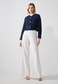 Cardigan court bleu marine structuré avec quatre poches avant et boutons dorés, associé à un pantalon large blanc et des sandales dorées à lanières.