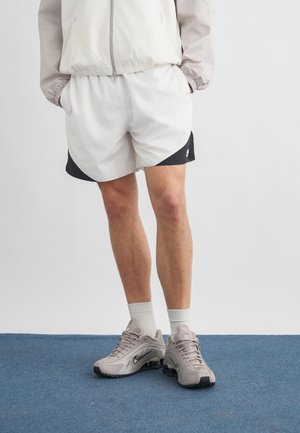 Person trägt beige-weiße Shorts, eine beige Jacke, weiße Sportsocken und hellgraue Nike-Laufschuhe und steht auf einem blauen Teppich.
