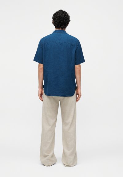 Άνδρας με σγουρά μαλλιά που φοράει μπλε κοντομάνικο πουκάμισο και μπεζ παντελόνια με φαρδιά πόδια, στέκεται γυρισμένος πλάτη σε απλό φόντο.
