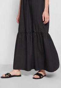 Robe maxi noire avec un ourlet froncé, faite d'un tissu lisse, portée avec des sandales noires plates à lanières tissées et au design minimal.