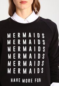 Donna che indossa una felpa nera con testo bianco ripetuto "MERMAIDS" e il testo aggiuntivo "HAVE MORE FUN" sottostante.