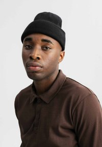 Zwarte gebreide beanie op het hoofd, gedragen in combinatie met een bruine polo met kraag en knopen. Glad textuur en neutrale tinten.