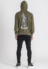 Felpa verde oliva con una stampa grafica sul retro, che presenta una figura alata e testo. Abbinata a jeans neri e sneaker.