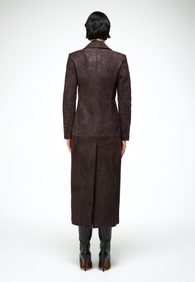 Personne vue de dos portant un long manteau en cuir marron foncé et des bottes à talons hauts noires sur un fond blanc.