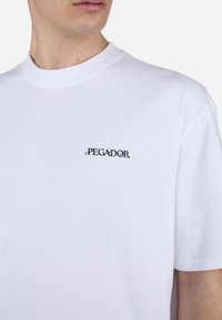 Persona che indossa una maglietta bianca con la scritta "PEGADOR" stampata in nero sulla parte superiore sinistra del petto, rivolta leggermente di lato.