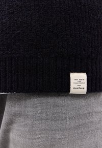 Granatowy sweter z teksturowanej dzianiny z prążkowanym dołem, z plecioną metką z napisem. Pod swetrem widać jasnoszary materiał.