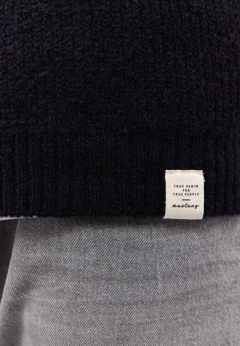 Granatowy sweter z teksturowanej dzianiny z prążkowanym dołem, z plecioną metką z napisem. Pod swetrem widać jasnoszary materiał.