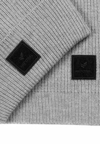 Grijze gebreide beanie met een geribd textuur. Heeft een zwarte rechthoekige tag met een ingekrast logo aan de voorkant en een kleinere tag aan de zijkant.