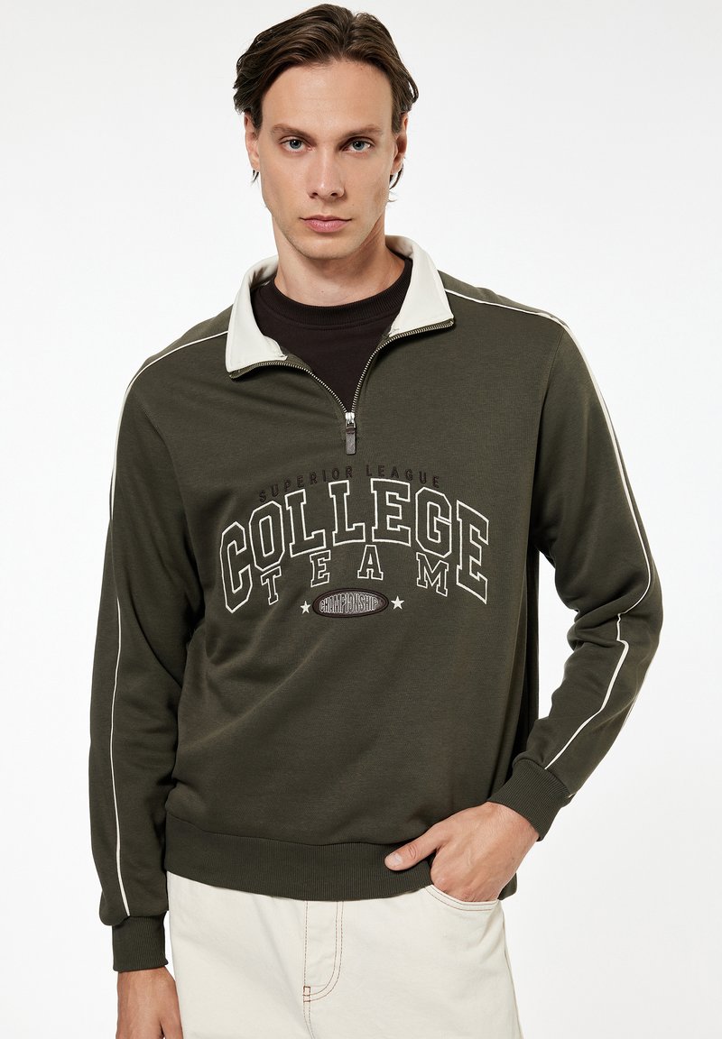 Man draagt een olijfgroene trui met halve rits en tekst "College Team", beige broek, staat met één hand in de zak, neutrale uitdrukking.