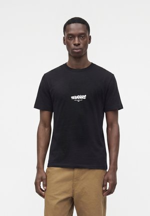 Ung mand iført en sort t-shirt med hvid tekst og beige bukser, stående mod en ensfarvet hvid baggrund.