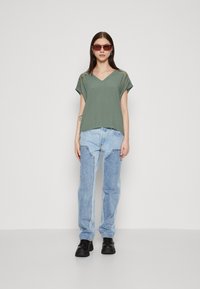 Top verde a scollo a V con maniche corte, abbinato a jeans oversize blu chiaro e scarpe con plateau nere, il tutto su uno sfondo neutro.