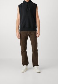 Mężczyzna w bezrękawniku w kolorze czarnym, ciemnobrązowych spodniach i białych trampkach stoi na tle jednolitego tła.