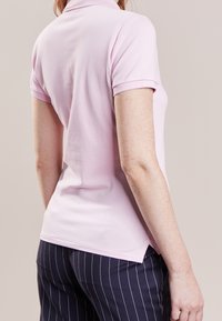 Ljusrosa polo skjorta med korta ärmar och klassisk krage, med en sidoflik vid fållen. Bärs tillsammans med marinblå byxor med rand.