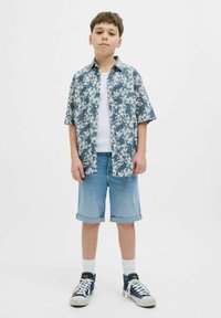 Camisa de flores azul de manga corta sobre una camiseta blanca, combinada con pantalones cortos de mezclilla azul claro y zapatillas náuticas con acentos blancos.