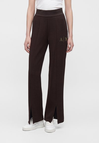 Pantalon noir à jambes larges avec fentes à l'avant de l'ourlet, logo doré "A|X" sur la cuisse, porté avec des baskets blanches et un haut blanc.