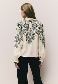 Bluză albă cu modele florale negre, mâneci lungi bufante cu mansete plisate și decolteu zimțat. Poartă cu pantaloni negri.