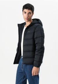 Black padded jacket with a hood, featuring a zip closure, elastic cuffs, and a small logo on the chest, paired with blue jeans.