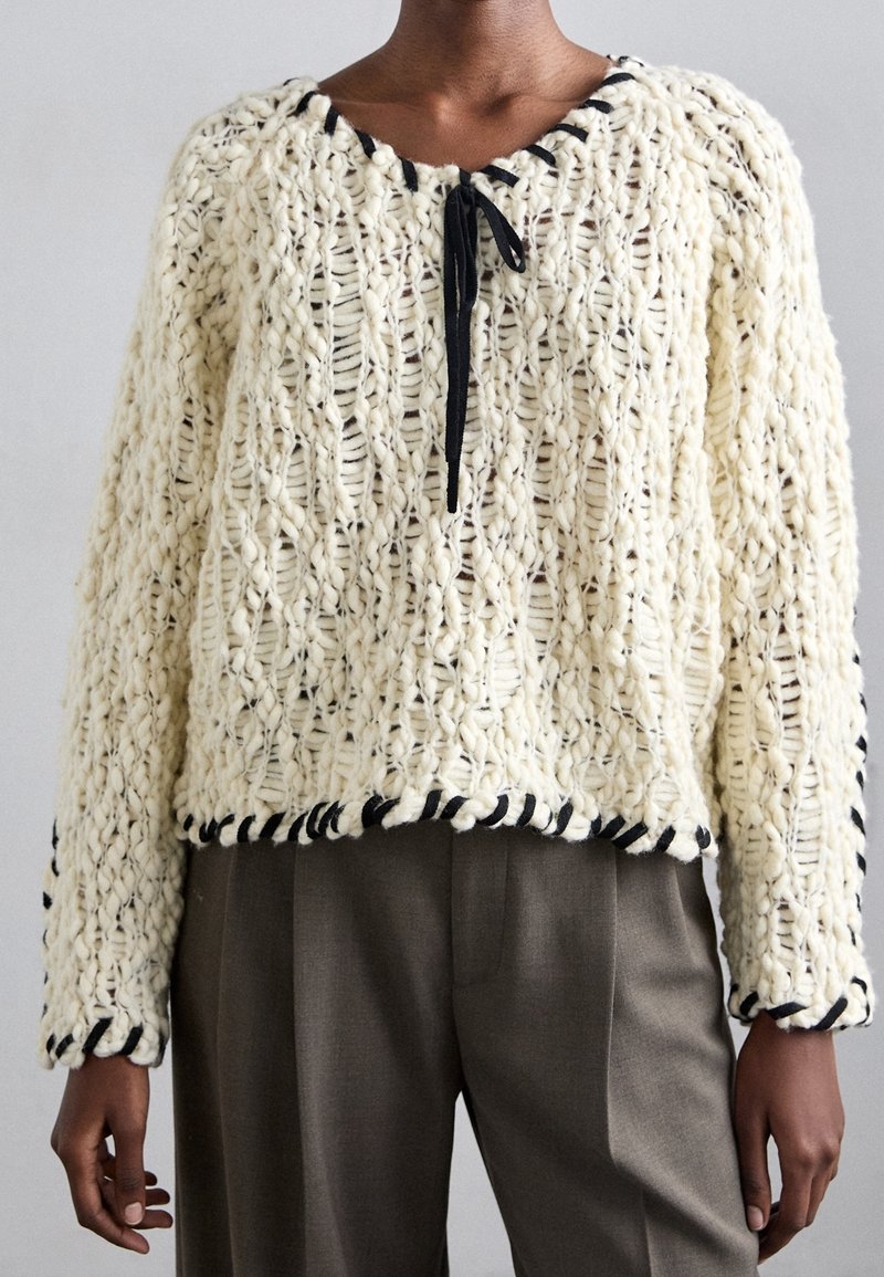 Pull en tricot crème épais avec bordure noire, col à nouer et manches longues porté avec un pantalon gris.