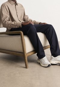 Persona sentada en una silla de cojín beige con marco de madera, vestida con una camisa de botones beige, pantalones oscuros y zapatillas blancas sobre un suelo neutro.