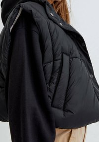 Černá vyp padded vesta s bezramenným designem, s texturovaným povrchem, patentkami a boční kapsou, vrstvená přes tmavý top.