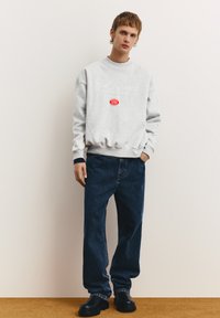 Grauer Sweatshirt mit dem Schriftzug "Pull Records" und einem roten Logo, kombiniert mit dunkelblauen, weit geschnittenen Jeans und schwarzen klobigen Schuhen.
