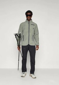 Άνδρας με γυαλιά ηλίου, γκρι φούτερ με φερμουάρ, μαύρο παντελόνι, λευκά αθλητικά παπούτσια, κρατώντας δύο μαύρους βατήρες σε μια ουδέτερη τοποθεσία.