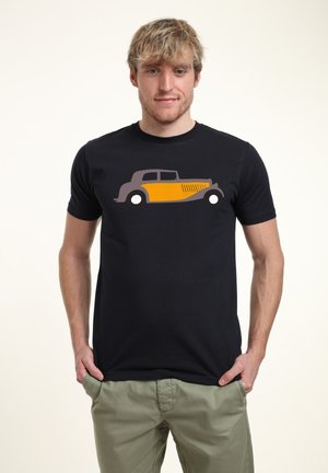Černé tričko s grafickým motivem vintage auta v šedé a žluté barvě. Modelka ho kombinuje se světle zelenými kalhotami. Jednoduchý design, krátké rukávy.