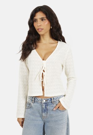 Vrouw met lang golvend haar, gekleed in een wit, textuurrijke blouse met strik aan de voorkant en lange mouwen, en lichtblauwe, hoog opgesneden jeans, met haar hand in de zak.