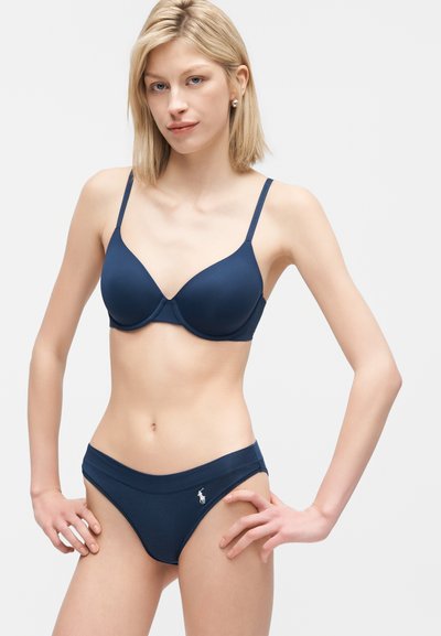 Ξανθιά γυναίκα με μπλε σκούρο σουτιέν και ταίριαστο εσώρουχο, όρθια με τα χέρια στη μέση μπροστά σε λευκό φόντο.