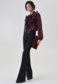 Doorzichtige bordeauxrode blouse met gerimpelde details, gecombineerd met zwarte hoge taille broek met gouden knopen. Houdt een bordeauxrode clutch vast.