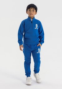 Blauwe sweatshirt en sweatpants set, met een halve rits kraag, een voorzak en witte cijfers "9" en "SAN FRI" gedrukt op beide stukken.