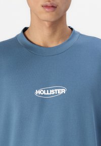 Kék pamut póló, kerek nyakkivágással, elöl fehér "HOLLISTER" logóval. Simább textúrájú, laza fazonnal.