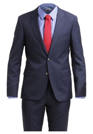 Homme marchant portant un costume bleu marine avec une chemise bleu clair, une cravate rouge et des chaussures marron sur fond blanc.