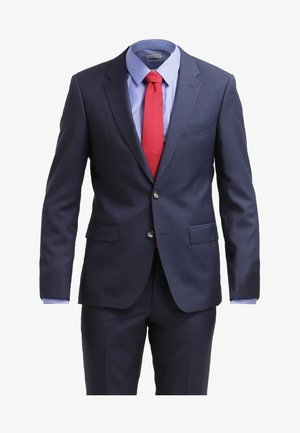 Hombre caminando vestido con un traje azul marino, camisa azul claro, corbata roja y zapatos marrones contra un fondo blanco.