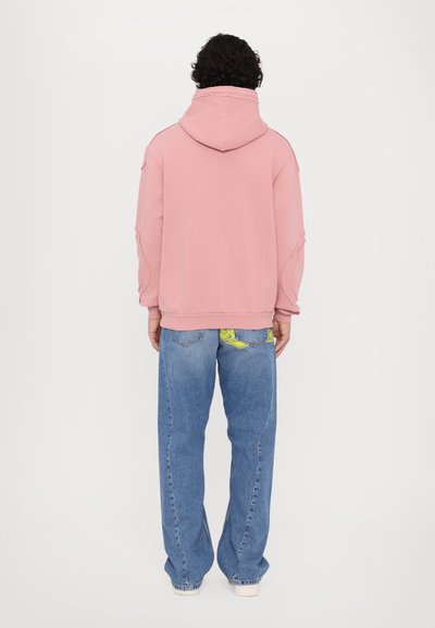 Rosa Kapuzenpullover mit lockerer Passform, der strukturierte Nähte aufweist. Kombiniert mit blauen weit geschnittenen Jeans, die eine gemusterte Innentasche zeigen.